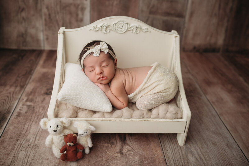 sleeping newborn baby girl laying on her tummy  bum up pose on top of tiny white wooden bed surrounded by 3 teddy bears with a brown wooden floor