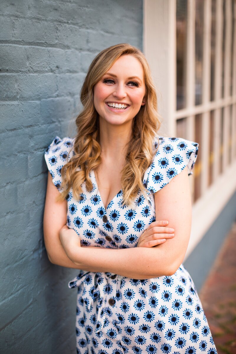 A blonde girl leans against a blue wall and smiles