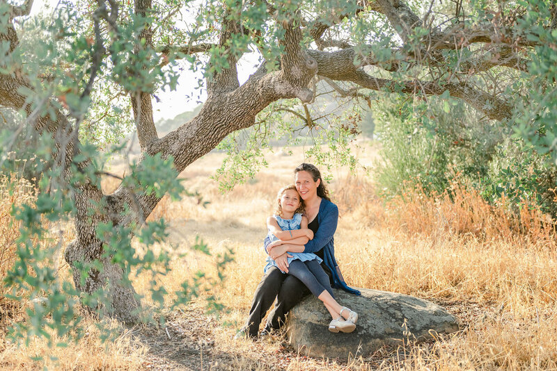 Mom in blue shirt holding her daughter in blue dress underneath an oak tree in the CA golden hills