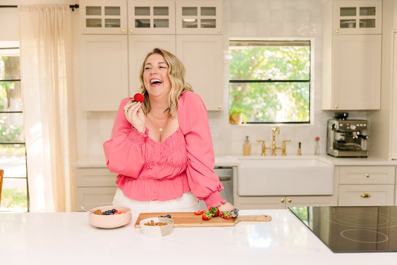 Mollie Mason in a bright pink top smiling and holding a strawberry standing at her kitchen island