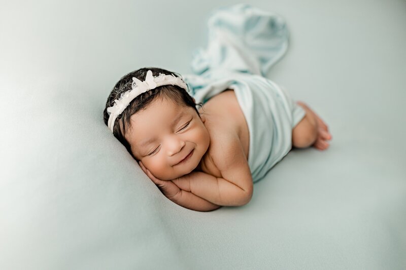Newborn baby girl lays on side with a bow on her head and a blue background