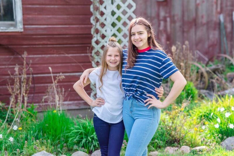 Two smiling girls posing together outdoors, one in a white shirt and the other in a striped blue shirt, standing by a rustic red building with a trellis.