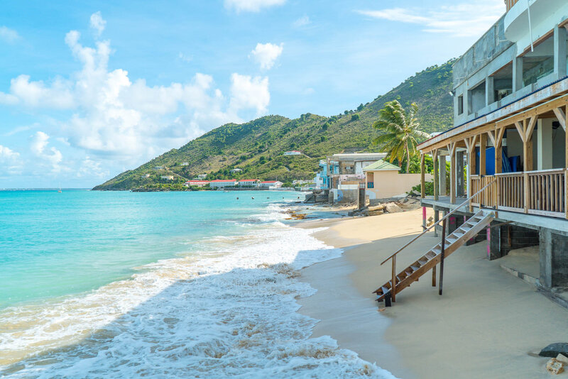 A scenic beach with turquoise water and gentle waves lapping at the sandy shore. Wooden buildings, including a two-story structure with a staircase, are on the right. Lush green hills rise in the background under a partly cloudy sky.