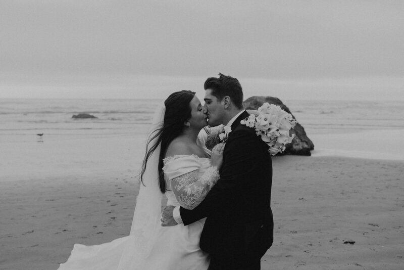 A couple kisses on Cannon Beach, dressed in wedding attire. The bride holds a bouquet, and the ocean waves and overcast sky create a serene backdrop.