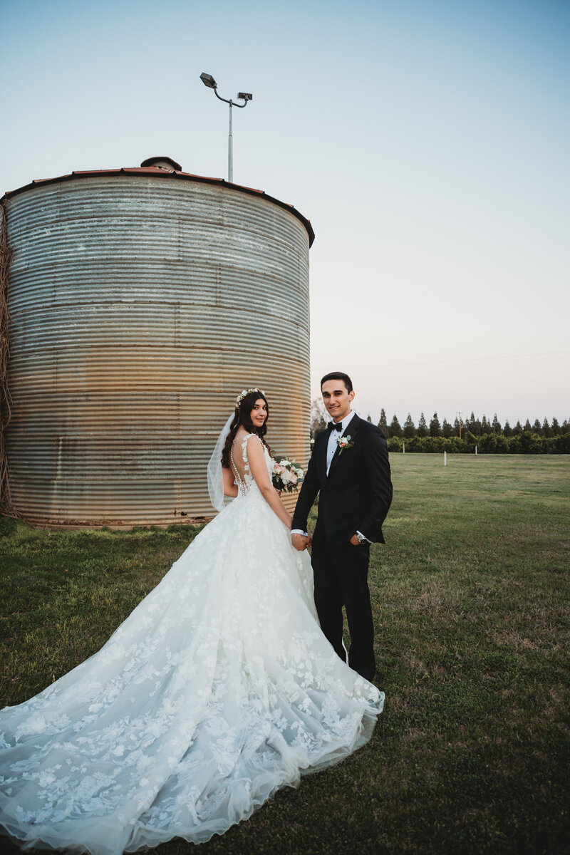 groom and bride with long flowing gown stand in front of silo