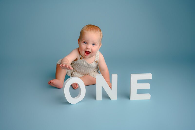 little boy sits in brown overalls playing with  ONE letters