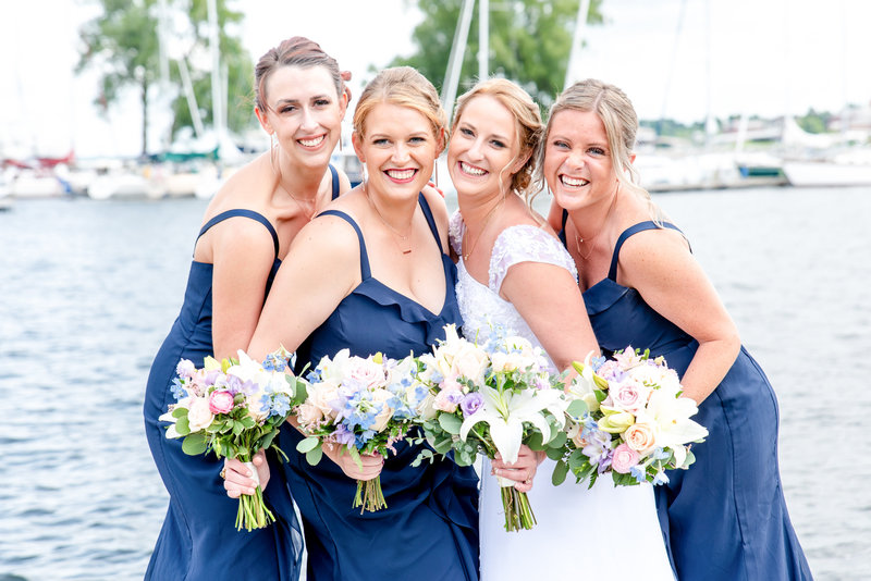 A bride with her bridesmaids in navy blue pose in front of sailboats