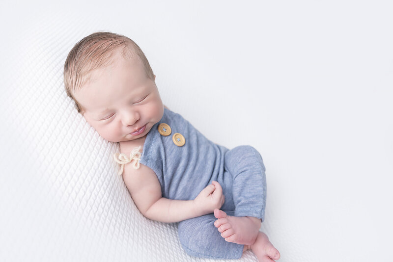 A sleeping newborn baby boy in blue overalls sleeps on a white pad