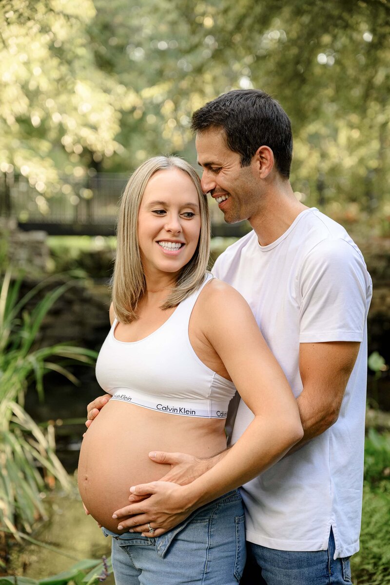 mother smiles as her husband stands behind her wrapping his arms around her touching her pregnant belly