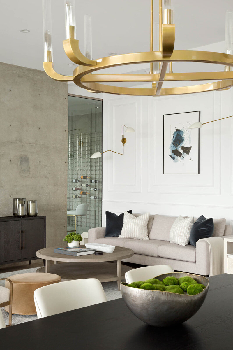 Rickridge project, in the foreground is a gold circular chandelier and a dark wooden dining table, in the background is the living room with an off-white sofa, grey wooden circular coffee table. The space is decorated with an abstract blue, black, and white painting, skinny golden wall sconces, greenery, and a cement wall