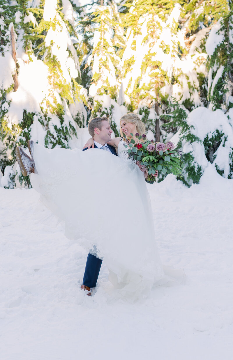 Groom picks up bride holds her and looks at her. Grouse mountain snowy photo and sunset in the back round. Couple in black and white attire for wedding day.