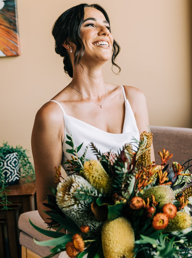the bride sitting by a window holding her flowers and laughing with joy