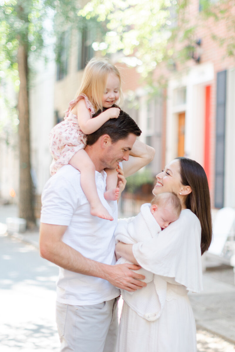 Mother holds sleeping newborn while dad and young daughter look on lovingly on Philadelphia street
