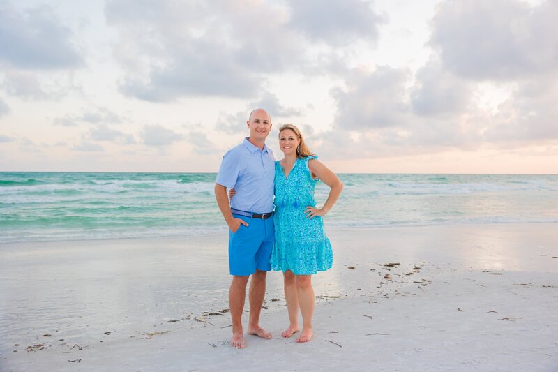 A husband and wife in blue outfits on the sand with Sarasota Bay behind them