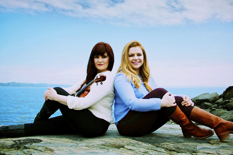 Music Duo photo Cassie and Maggie sitting on rocks knees bent ocean behind them