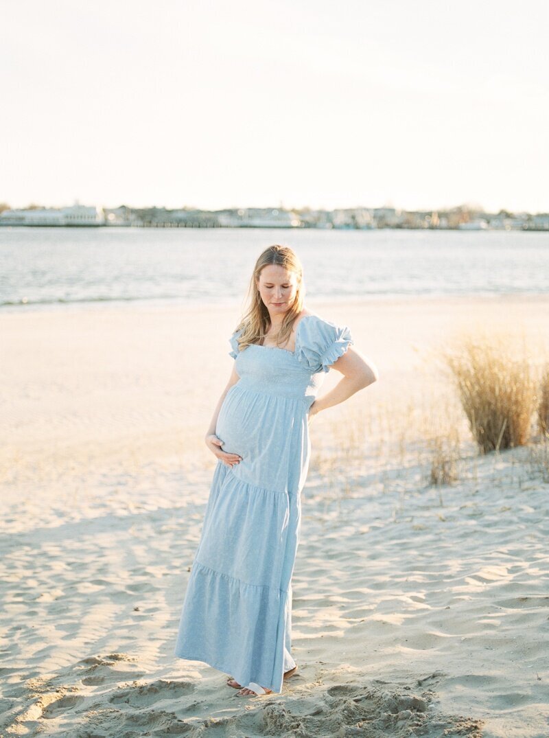 A woman stands on the beach during her maternity photos in NJ. She is wearing a blue dress