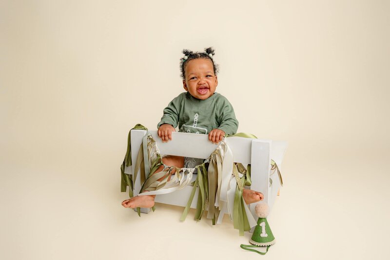 litte one year old girl sits on white bench dressed in green with a one year old birthday banner on the bench