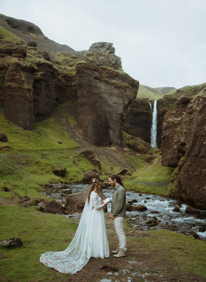 bride and groom are reading vows to one another with a waterfall in the background. they are standing by a river and the groom as his hands interlocked while the bride reads her vows.
