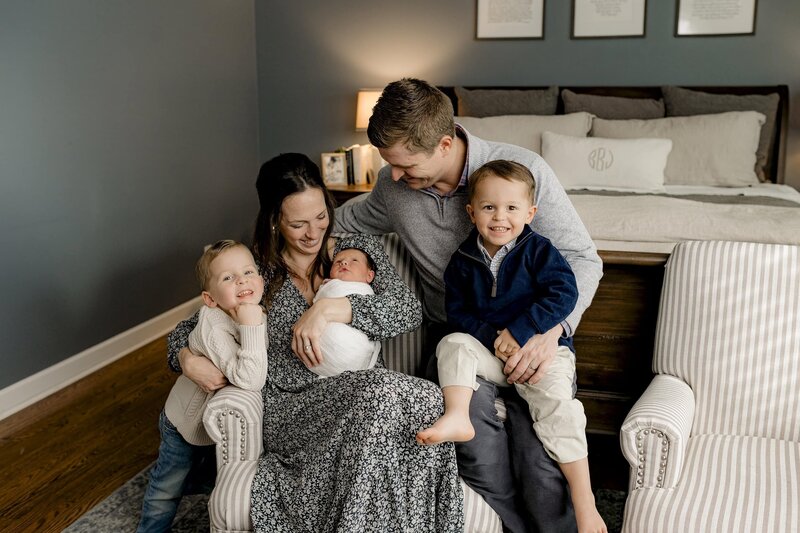 mother and father sit together while Mom holds the newborn baby and each of their sons is beside them with everyone looking down at the newborn and the two sons looking towards the camera