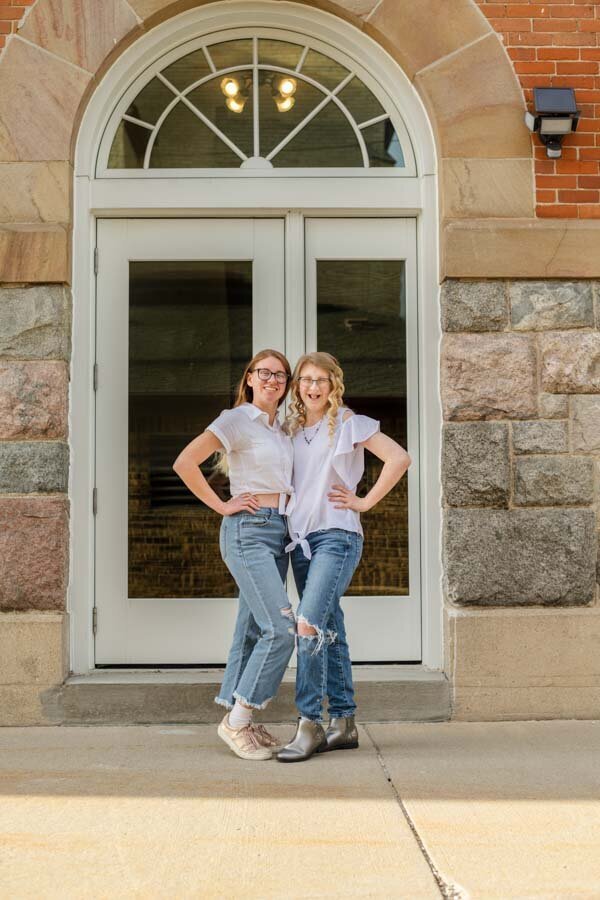 Two young women in white t-shirts and jeans, standing confidently with hands on hips in front of an arched doorway.