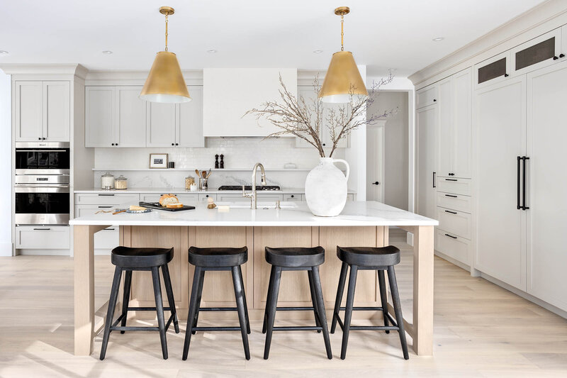 Sinclair project, a large white marble kitchen island, with 4 black wooden stools below. The island frame and floors are a light wood. The kitchen is all white with gold hanging pendent lights above the island