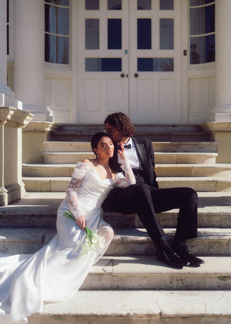 Bride and groom sitting on elegant stone steps in soft sunlight, captured in a romantic, cinematic style by a luxury documentary wedding photographer UK.