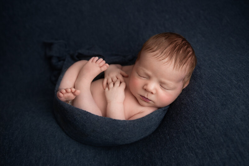 Hampton London studio photograph of a sleeping newborn baby boy wrapped in & laying on a navy blue fabric.