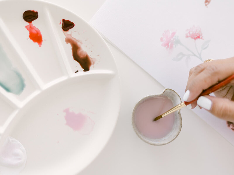 A brush dipped into a watercolor palette beside a partially drawn pink watercolor flower design on paper