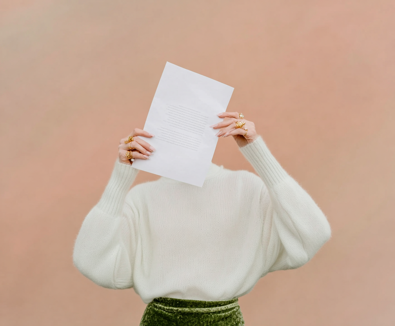 Woman in sculptural white sweater holding a letter in front of her face, set against a blush backdrop. Editorial image used on Jasmina Kimova’s contact page.