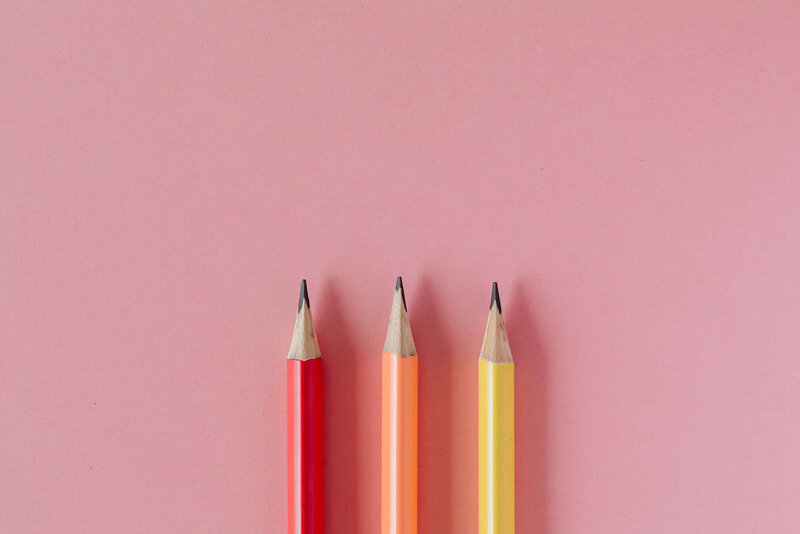 Three sharpened pencils in red, orange, and yellow are neatly aligned on a pink background.