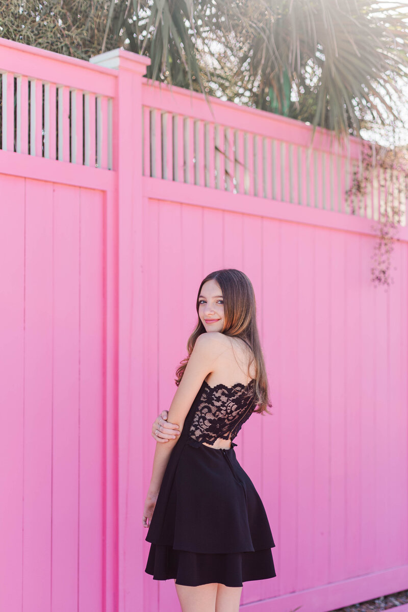 A shy sixteen year old poses in front of a pink fence in a black lace dress in Galveston Texas