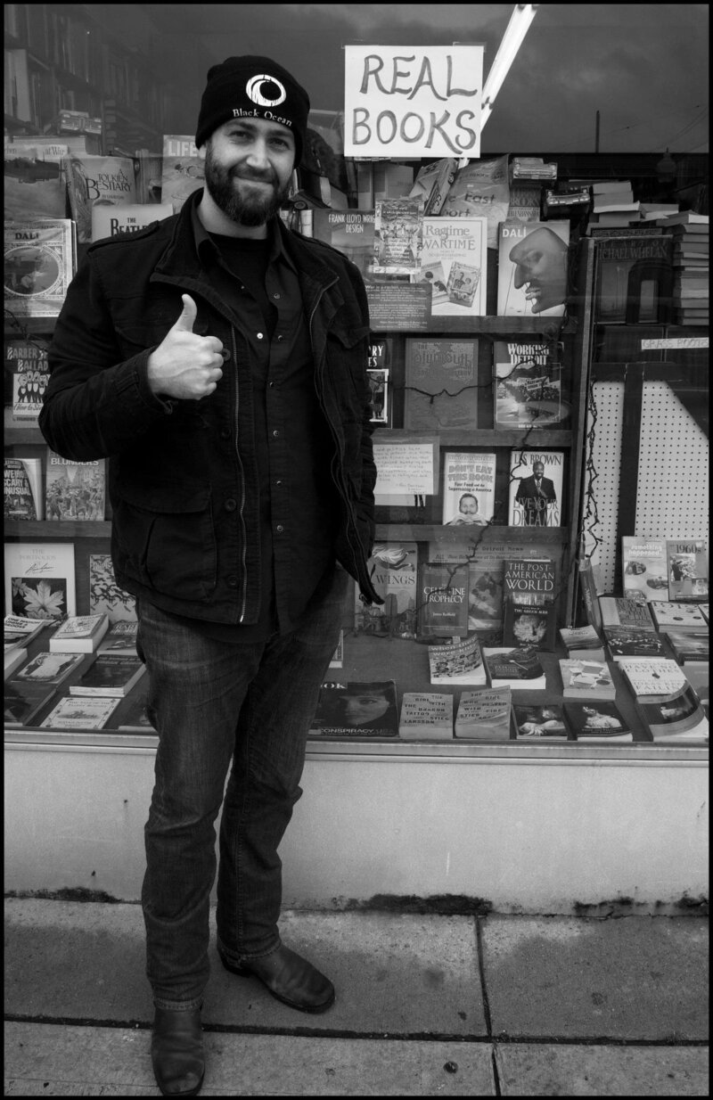 Janaka Stucky standing in front of a bookstore window with a sign advertising "Real Books"