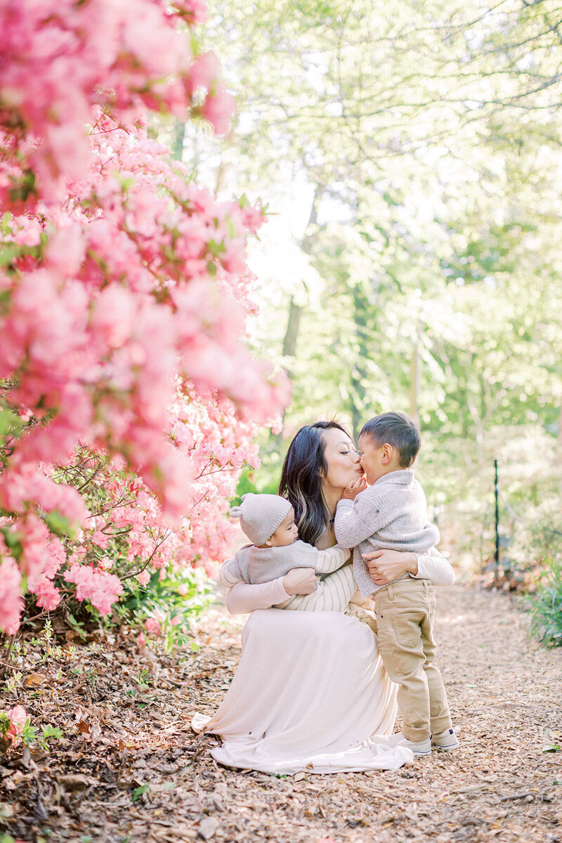 A mother crouches down and kisses her toddler son while holding her baby near some pink azaleas at Brookside Gardens