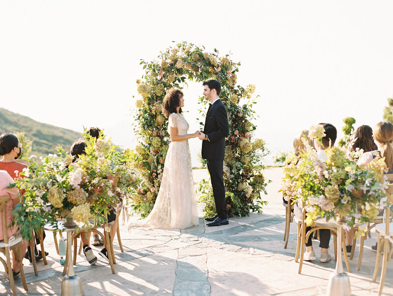 Couple getting married during their ceremony. They are holding hands standing in front of a super lush floral arch