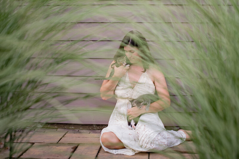 Woman in cream lace dress holding kittens with tall green grass in front