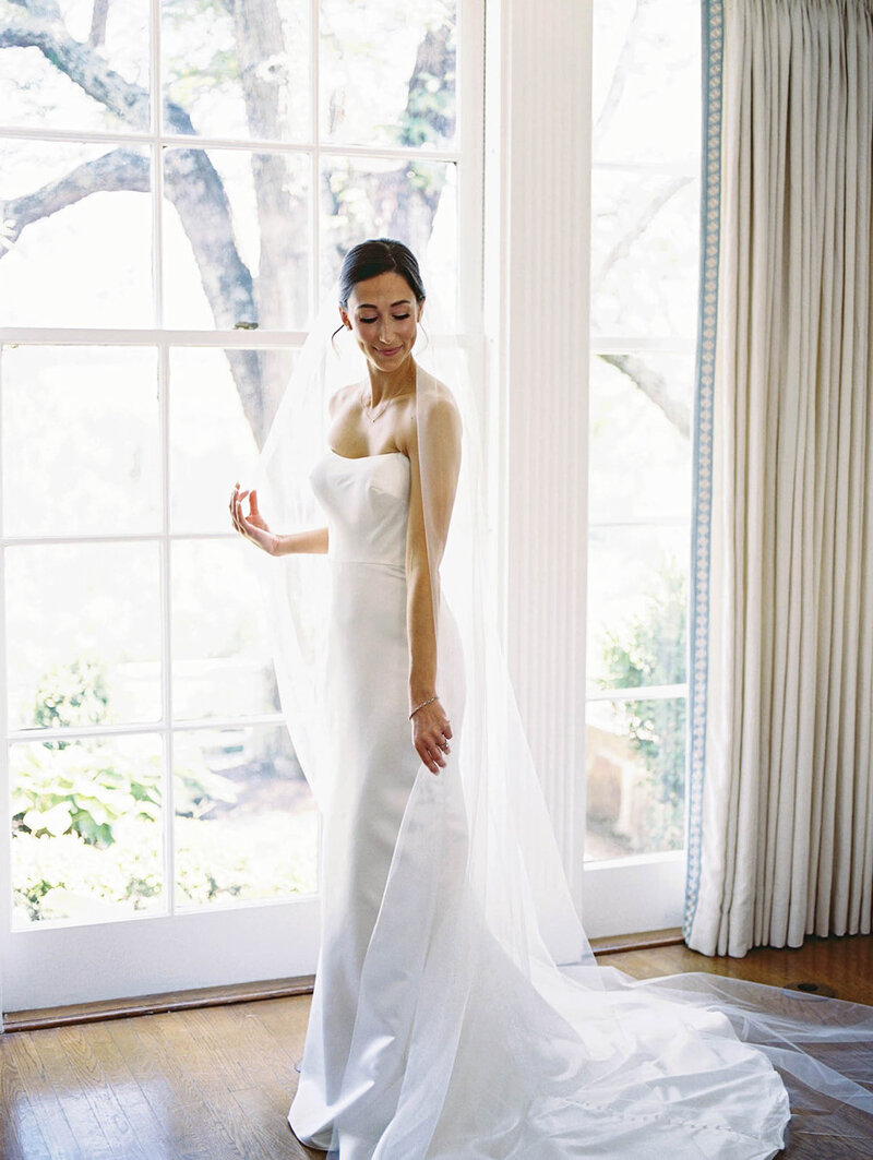 Bride by a floorlength window looking out as she holds hands together and her veil drapes the floor.