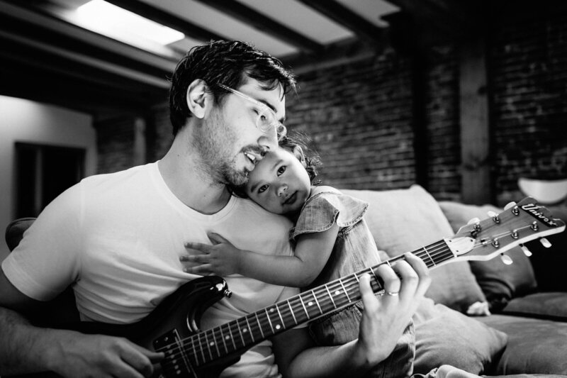monochromatic image of father tenderly holding toddler daughter while he plays electric guitar from an in-home photo session.