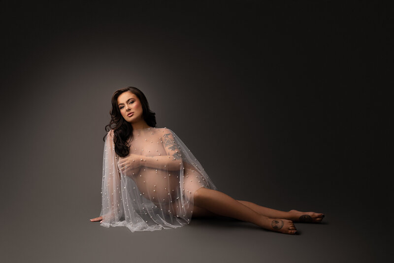 Woman poses for a Philadelphia Main Line fine art maternity photoshoot. Woman is sitting on the ground. She is bare and draped in a pearl-beaded organza sheath. She is sitting with her legs to the side, weight resting on her left arm and her right arm is covering her breasts. She is looking at the camera with a serious expression. Captured by best Philadelphia maternity photographer Katie Marshall.