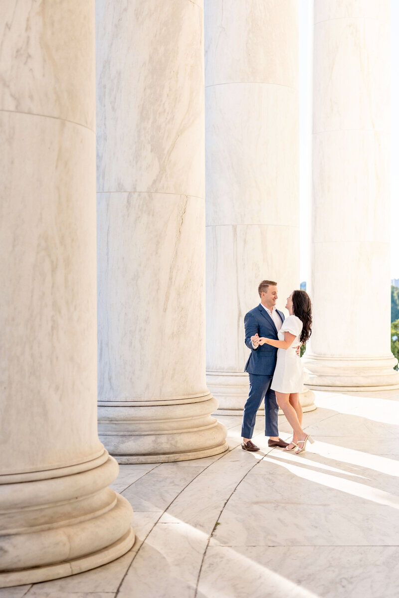A couple stands closely together, smiling at each other, surrounded by large marble columns. The man is in a blue suit, and the woman is in a white dress. The scene is sunlit, creating a serene and romantic atmosphere.