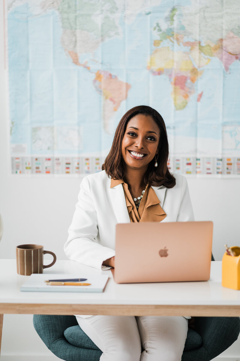 A smiling woman in a business suit sits at her desk with a laptop, ready to assist clients as a destination wedding planner.