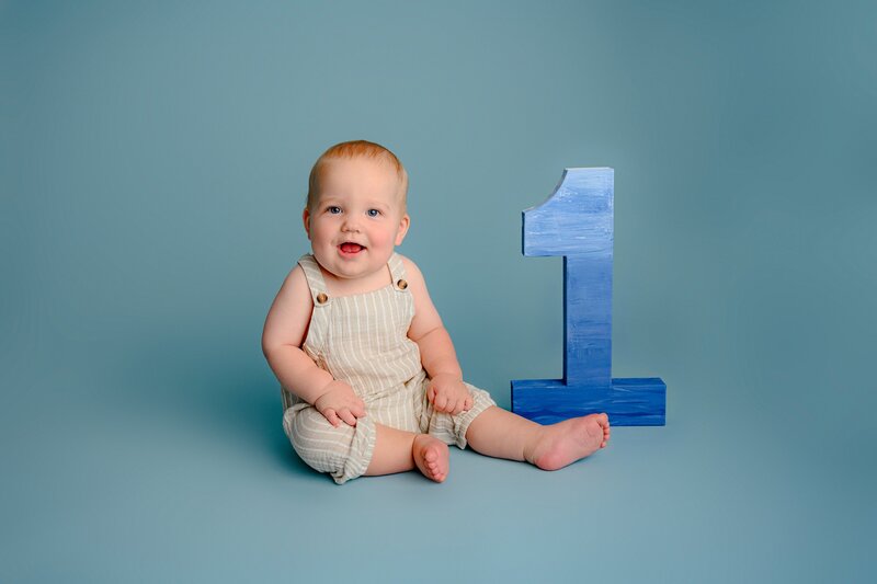 little one year boy sits next to a number one prop dressed in a brown overalls and blue background