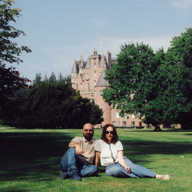 A husband and wife photography duo sit together on the grass in front of a historic castle, relaxed and smiling in the afternoon light.