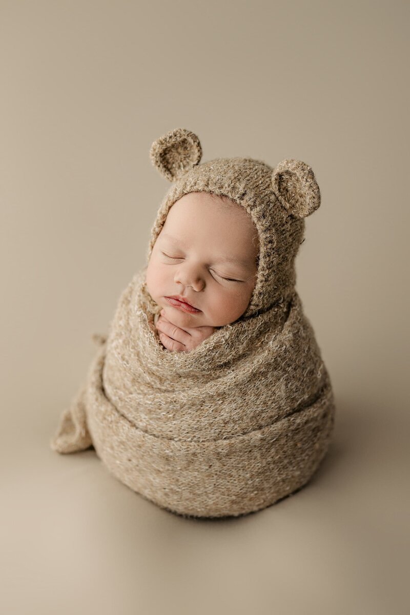 Infant boy is wearing a bear bonnet with ears and a matching neutral colored wrap sitting upright during his Studio newborn session