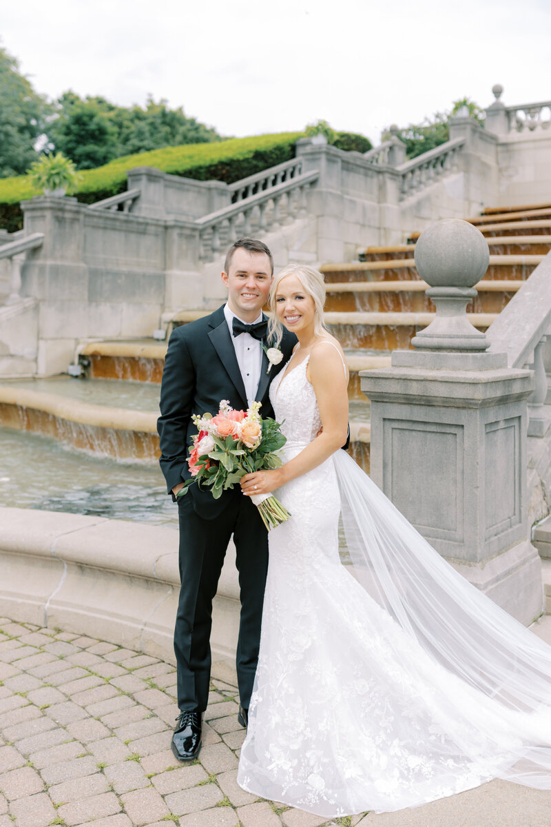 A couple smile towards the camera in a classic portrait in front of the fountain at Ault Park