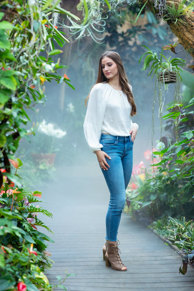 Senior girl posing looking to the left wearing jeans and heels with w light top standing in the middle of a greenery path