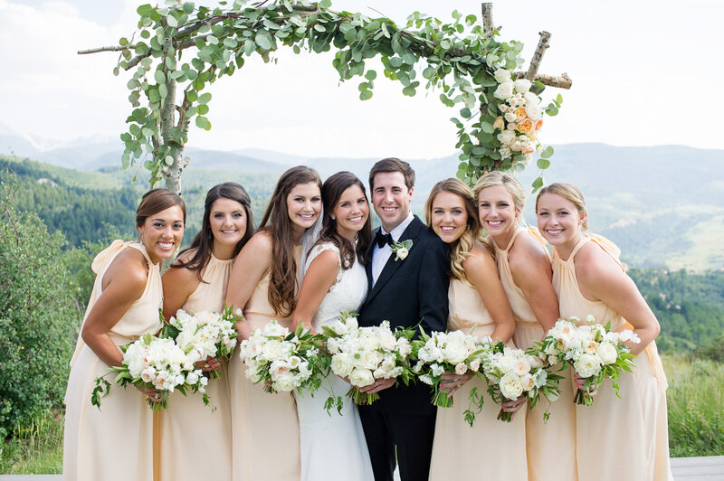 Testimonial photo, bride and groom with 6 bridesmaids surrounding them smile at the camera, behind them is a wooden arch decorated with foliage