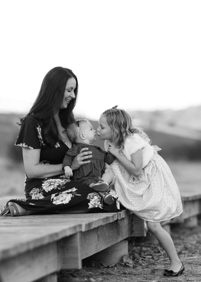 mom and daughters smiling and kissing on bridge