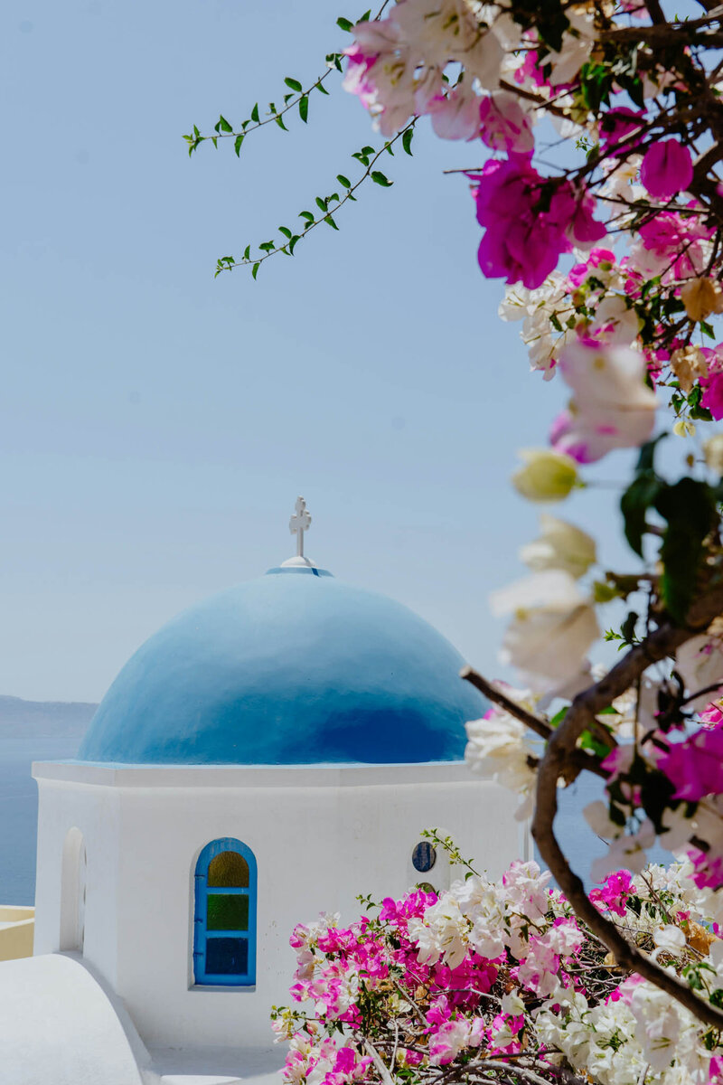 A white building with a blue dome and cross, framed by vibrant pink and white bougainvillea flowers. The background shows a clear sky and distant sea, evoking a serene Mediterranean vibe.