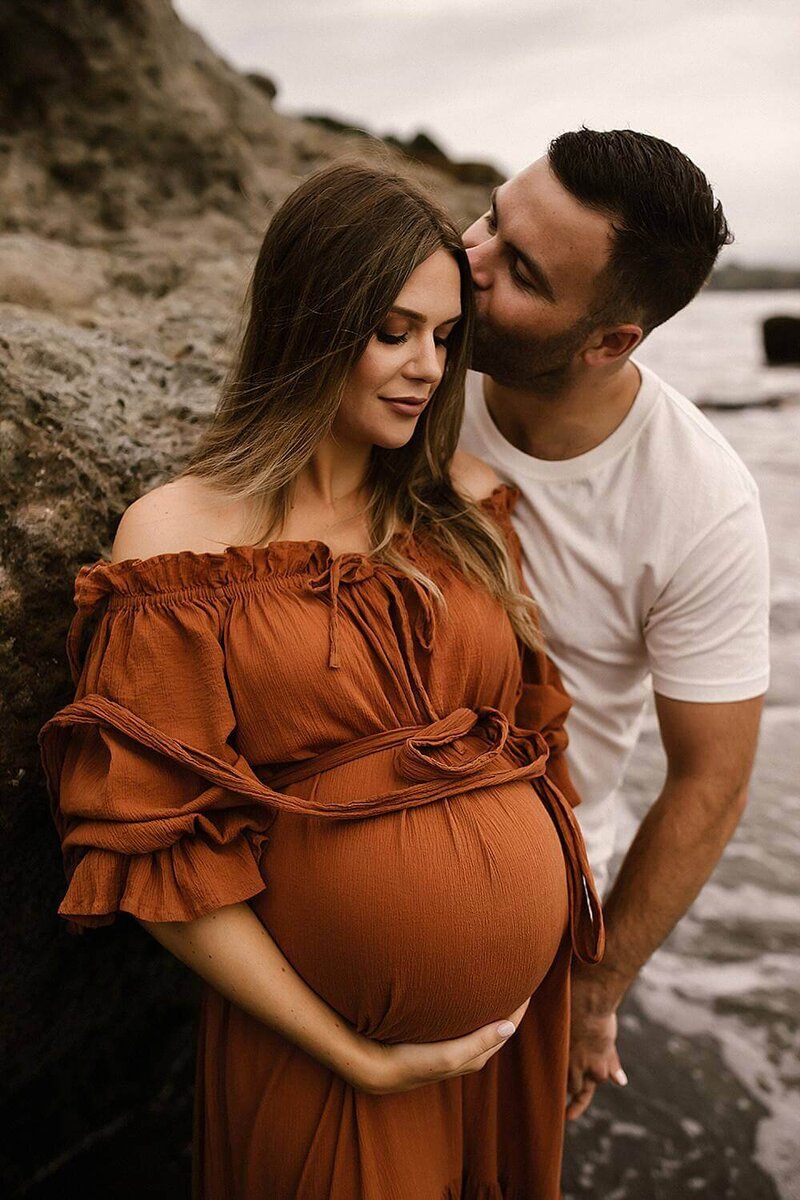 a husband and wife wearing white and mustard during their maternity photoshoot at a local new plymouth beach with aimee kelly photography