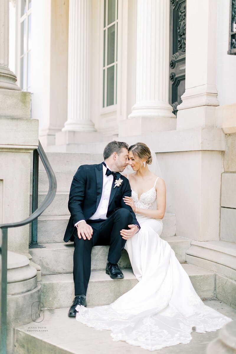 Bride and Groom sit on marble stairs outside. They have their foreheads touching.